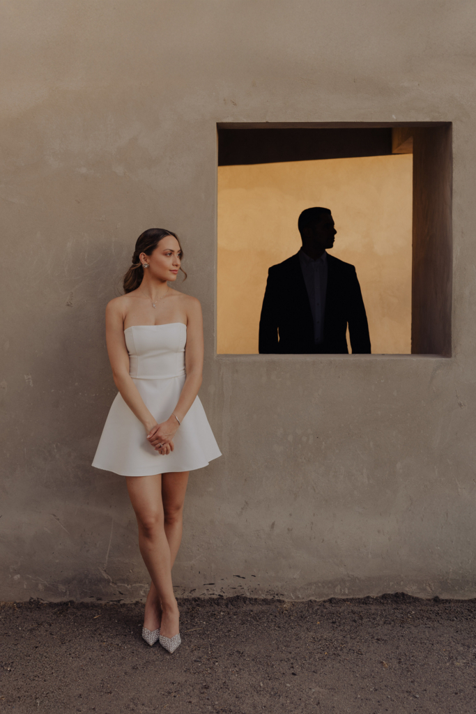 This is a color portrait of a woman in a short white dress standing against a textured wall. She looks to the side with her hands gently clasped, while a rectangular window beside her frames the silhouette of a man in shadow. The composition contrasts soft, warm light on her figure with the dark, graphic shape inside the window.