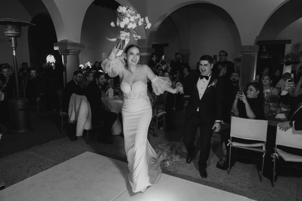 This is a black-and-white photograph of a bride running joyfully through a reception space, holding her bouquet high in the air. The groom follows behind her, smiling, while guests watch and capture the moment on their phones. The scene is lively and energetic, with movement and celebration filling the frame.