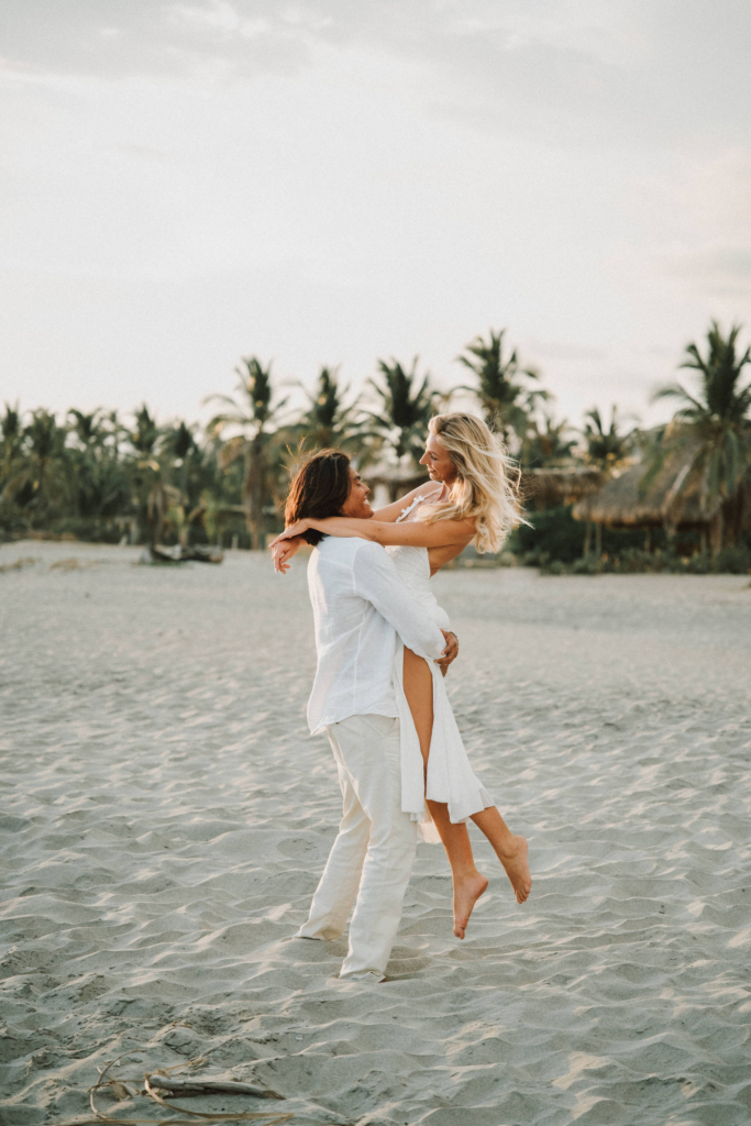 A couple dressed in white share playful, intimate moments on a quiet sandy beach lined with palm trees. The images feel light, romantic, and carefree, with soft natural light and a relaxed tropical atmosphere.