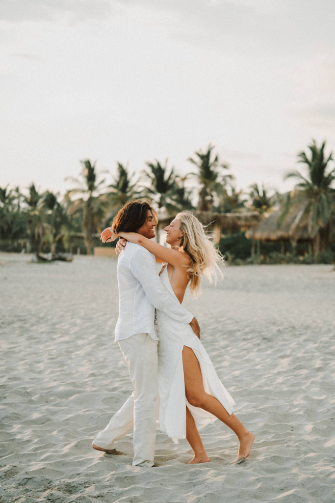 A couple dressed in white share playful, intimate moments on a quiet sandy beach lined with palm trees. The images feel light, romantic, and carefree, with soft natural light and a relaxed tropical atmosphere.