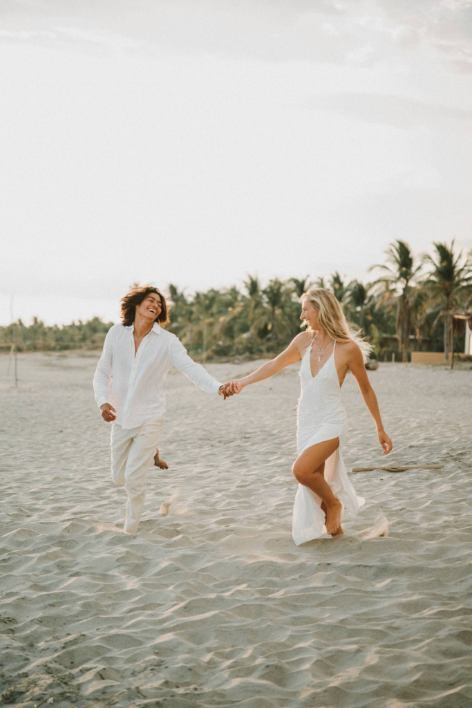 A couple dressed in white share playful, intimate moments on a quiet sandy beach lined with palm trees. The images feel light, romantic, and carefree, with soft natural light and a relaxed tropical atmosphere.