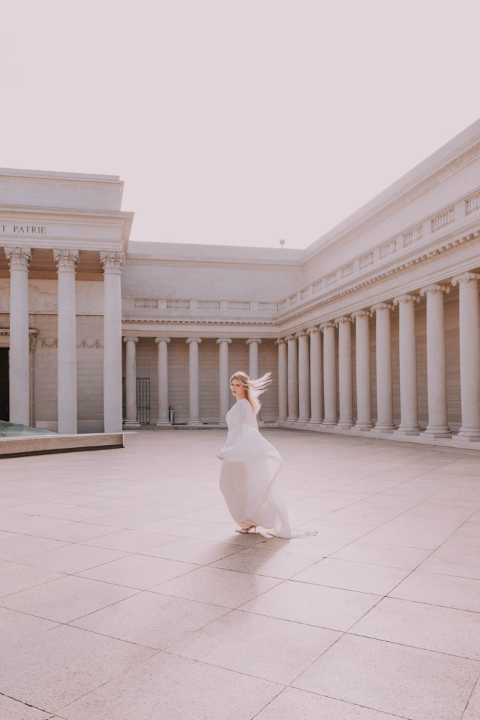 A woman in a flowing white dress walks across a bright, open courtyard surrounded by classical columns.
Her hair and dress move gently in the light, creating a soft, airy feeling against the pale stone architecture.