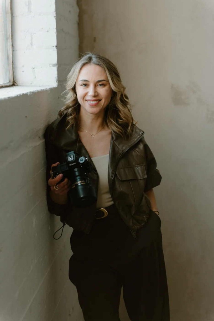 A portrait of wedding photographer Monika Grzelak standing by a window, holding a camera and looking at the viewer in soft natural light.