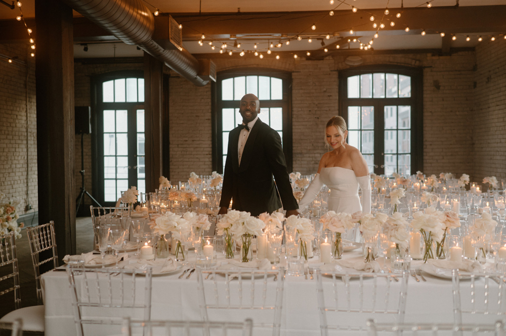 A couple walks hand in hand through a reception space filled with long tables, candles, and white floral arrangements.