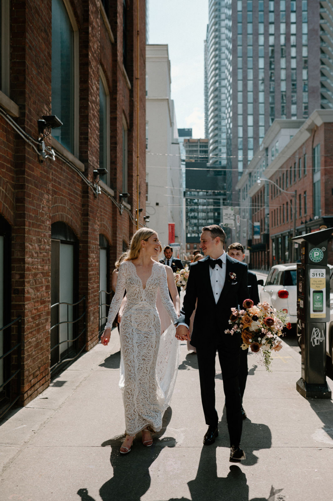A bride and groom walk hand in hand down a city sidewalk in daylight, surrounded by tall buildings and urban details. They look at each other with relaxed smiles, carrying a bouquet of warm-toned flowers. The scene blends wedding elegance with a casual, everyday city atmosphere.