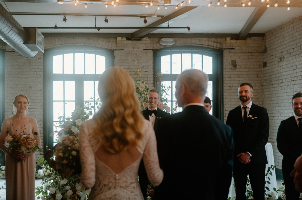A bride walks down the aisle with an older man, approaching the groom as guests look on inside a bright indoor ceremony space.