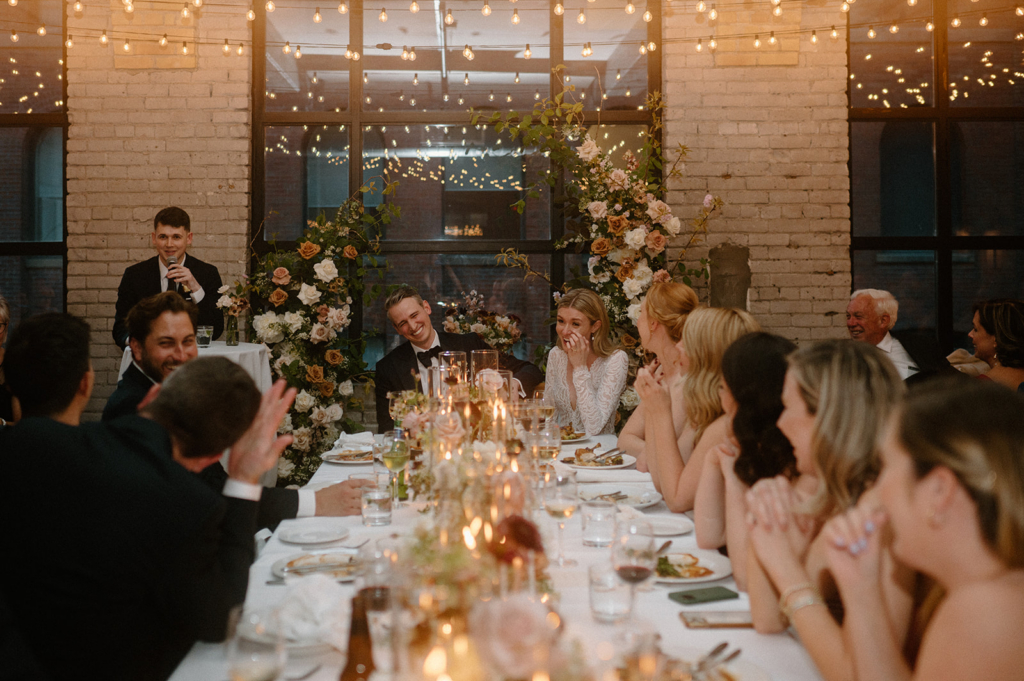 A bride and groom stand close together between candlelit tables, surrounded by flowers in a softly lit reception setting.