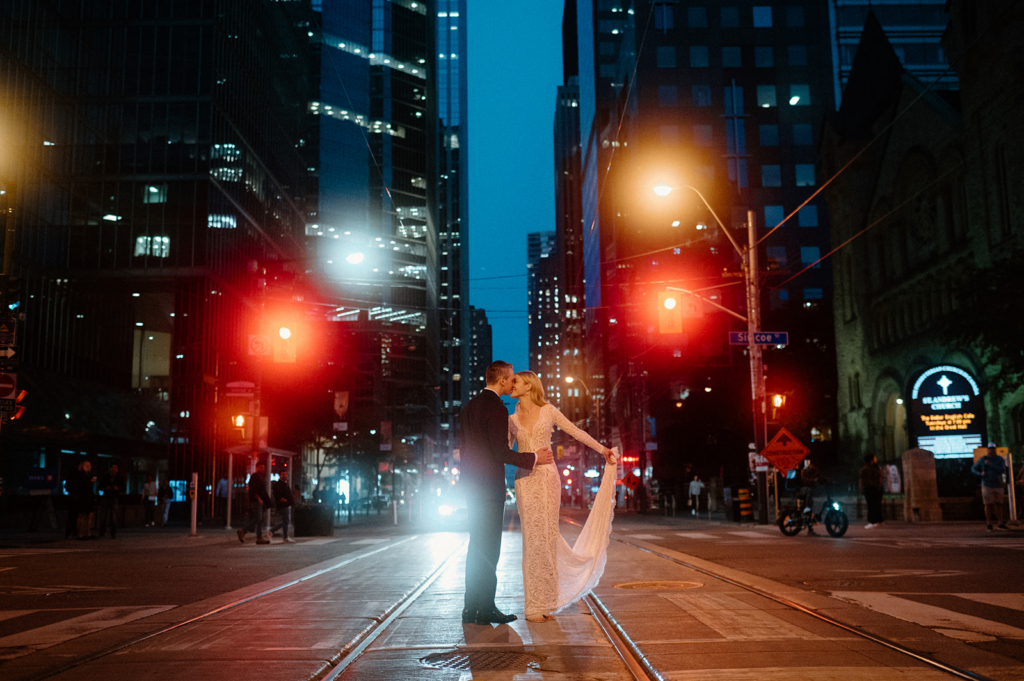 A couple stands in the middle of a city street at night, facing each other and about to kiss, surrounded by tall buildings and glowing traffic lights.