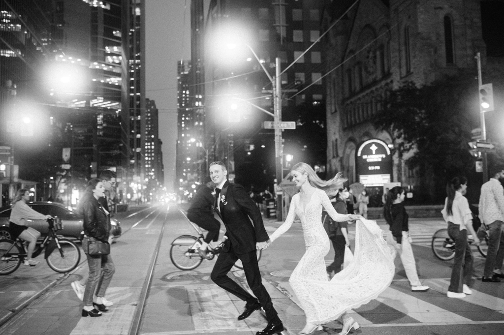A bride and groom run hand in hand across a city street at night, surrounded by glowing streetlights and passing pedestrians. The bride’s dress flows behind her as they move, creating a sense of motion and spontaneity. The black-and-white image captures an energetic, cinematic moment within the rhythm of the urban environment.