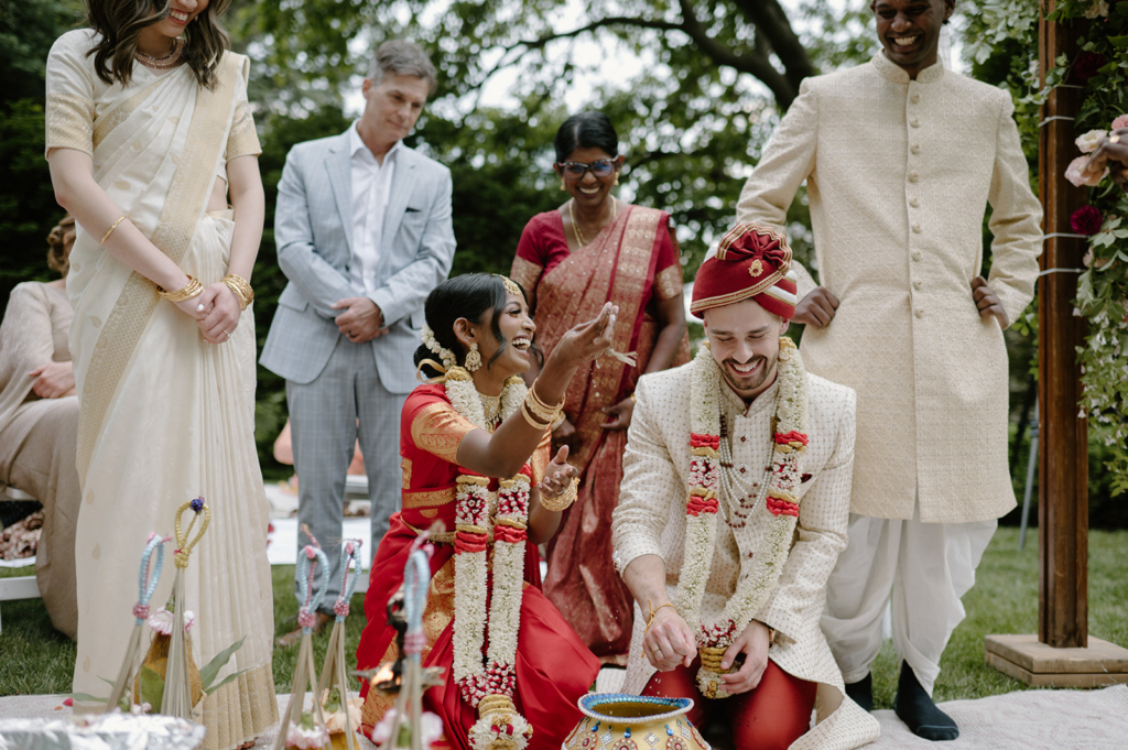 A couple takes part in a traditional ceremony outdoors, smiling and laughing as family members stand around them.