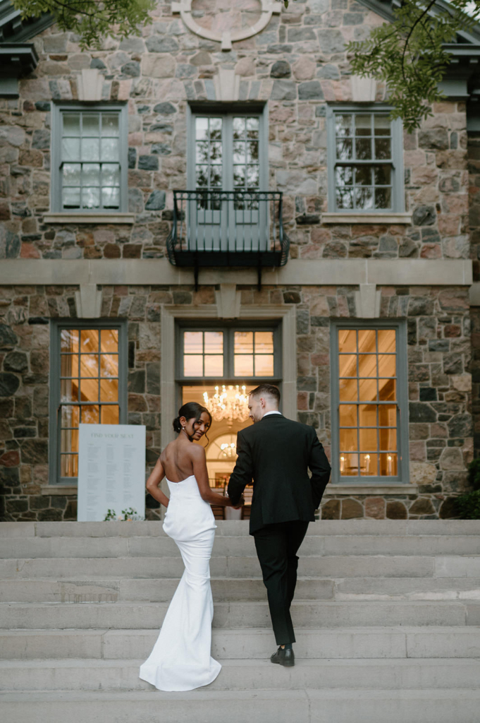 A bride and groom walk up stone steps toward a large, elegant building with lit windows behind them. The bride glances back over her shoulder, creating a quiet, cinematic moment as they enter the venue together. The setting feels intimate and refined, with soft evening light enhancing the scene.