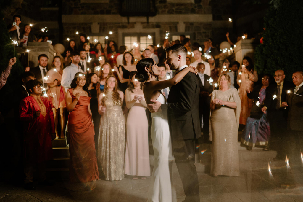 A bride and groom share a slow dance at night, surrounded by a crowd of guests holding sparklers that glow warmly in the dark. The couple stands at the center, embraced and focused on each other, while the guests form a luminous circle around them. The scene feels celebratory and intimate, blending movement, light, and emotion in a single moment.