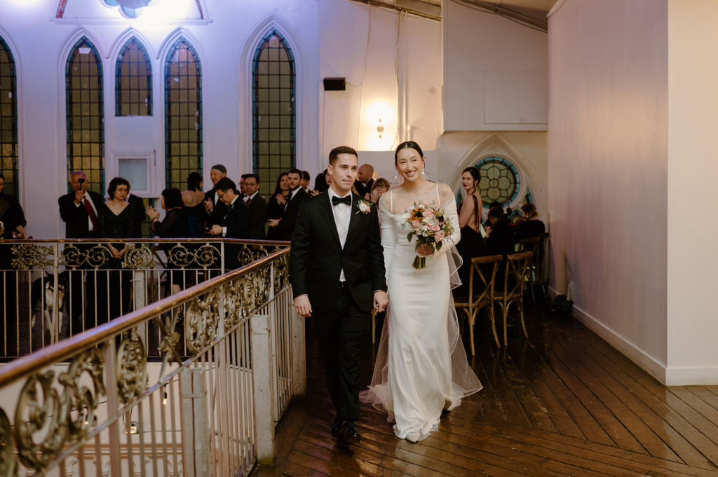 A bride and groom walk together through an indoor venue, smiling as guests gather and watch them from the background.