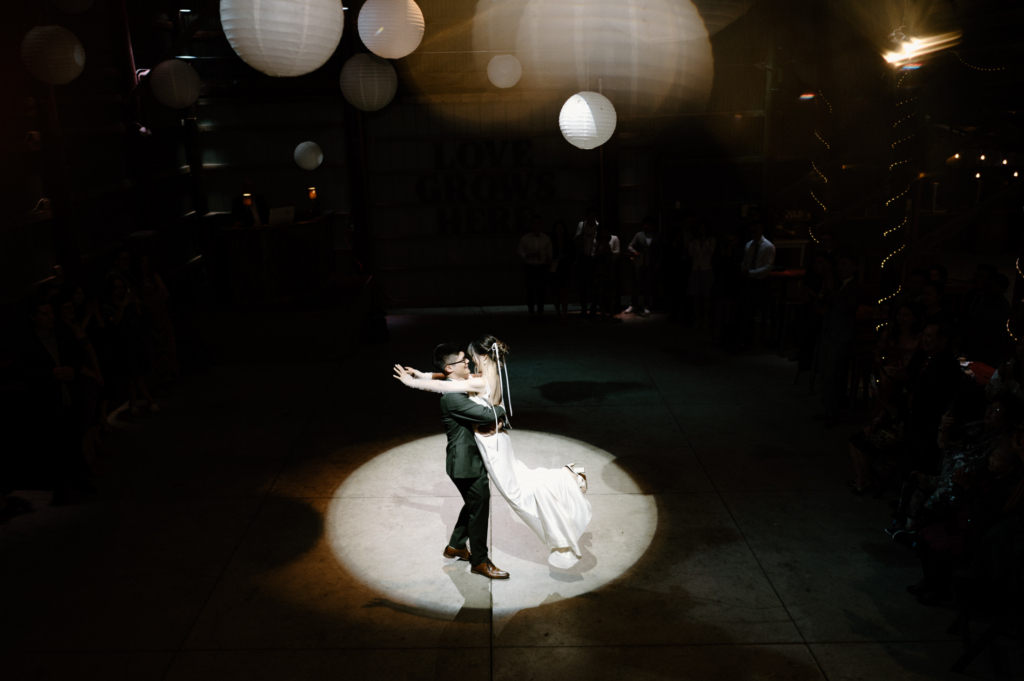 A bride and groom share their first dance under a spotlight, surrounded by darkness and hanging lights above.