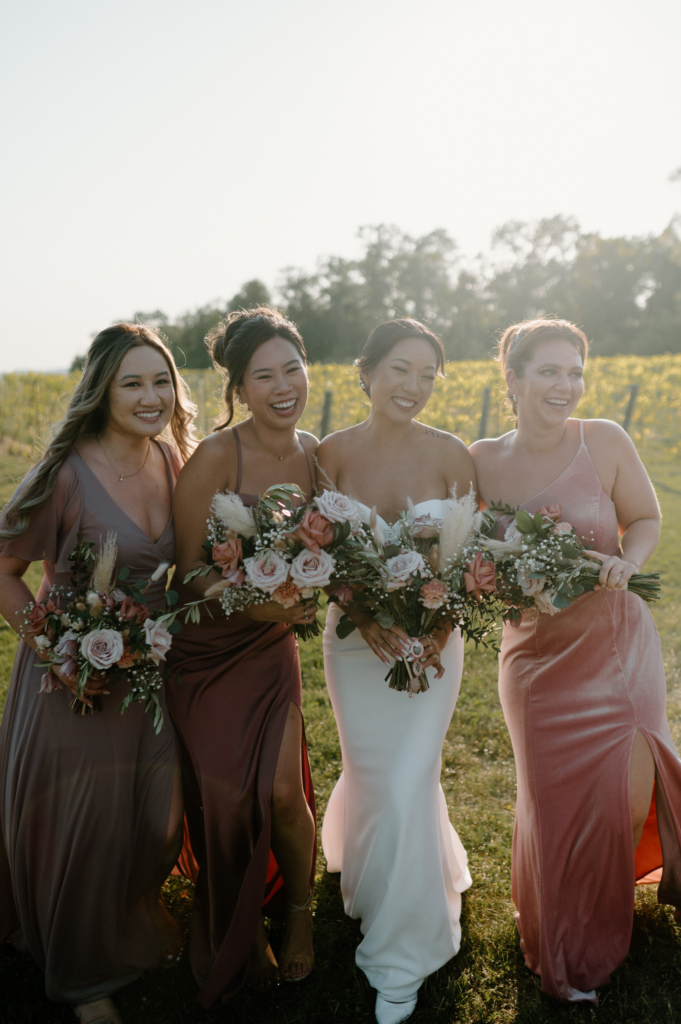 A bride stands with three bridesmaids outdoors, all holding bouquets and smiling together in soft natural light.
