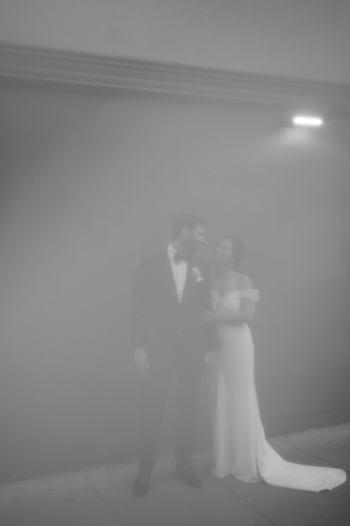 A bride and groom stand close together in a foggy street at night, partially obscured by mist and soft light.