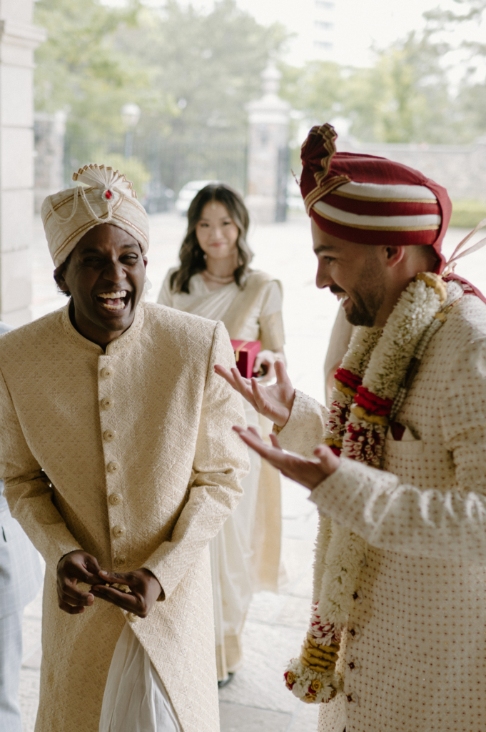 A groom and another man in traditional attire laugh together during a wedding moment, while a woman stands behind them holding a gift.
