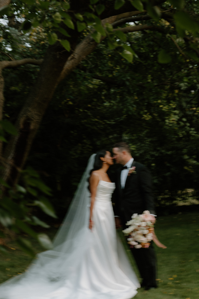 A bride and groom stand close together under trees, softly blurred in motion as they lean in for a kiss in a green outdoor setting.