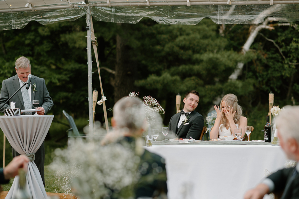 A groom watches the bride with a smile as she reacts emotionally during a wedding speech, while a guest speaks at a microphone nearby.