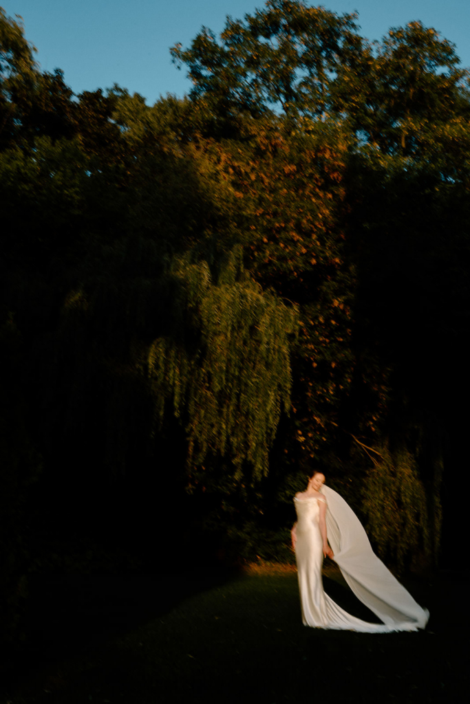 A bride stands alone outdoors at sunset, her long veil flowing behind her against a dark, tree-filled background.