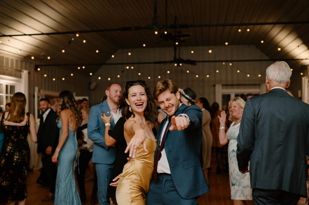 A man and woman dance together at a lively wedding reception, smiling and pointing toward the camera as guests gather around them.