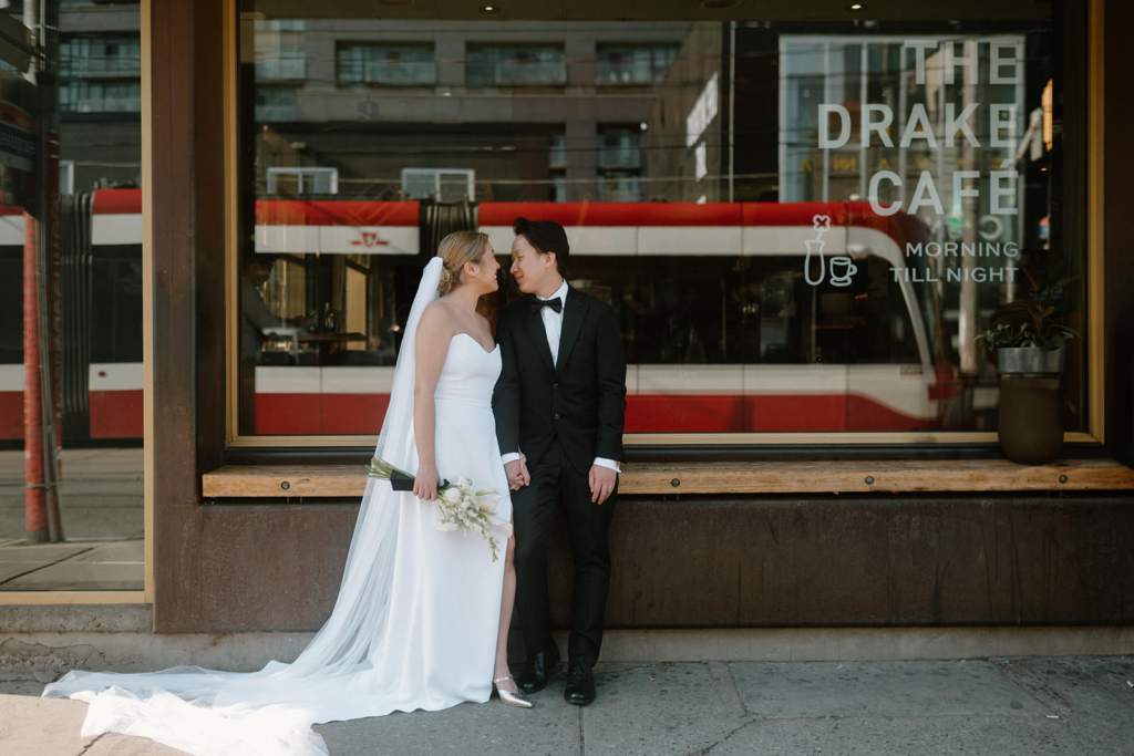 A bride and groom stand outside a café window, holding hands and smiling at each other, with a red streetcar reflected behind them.