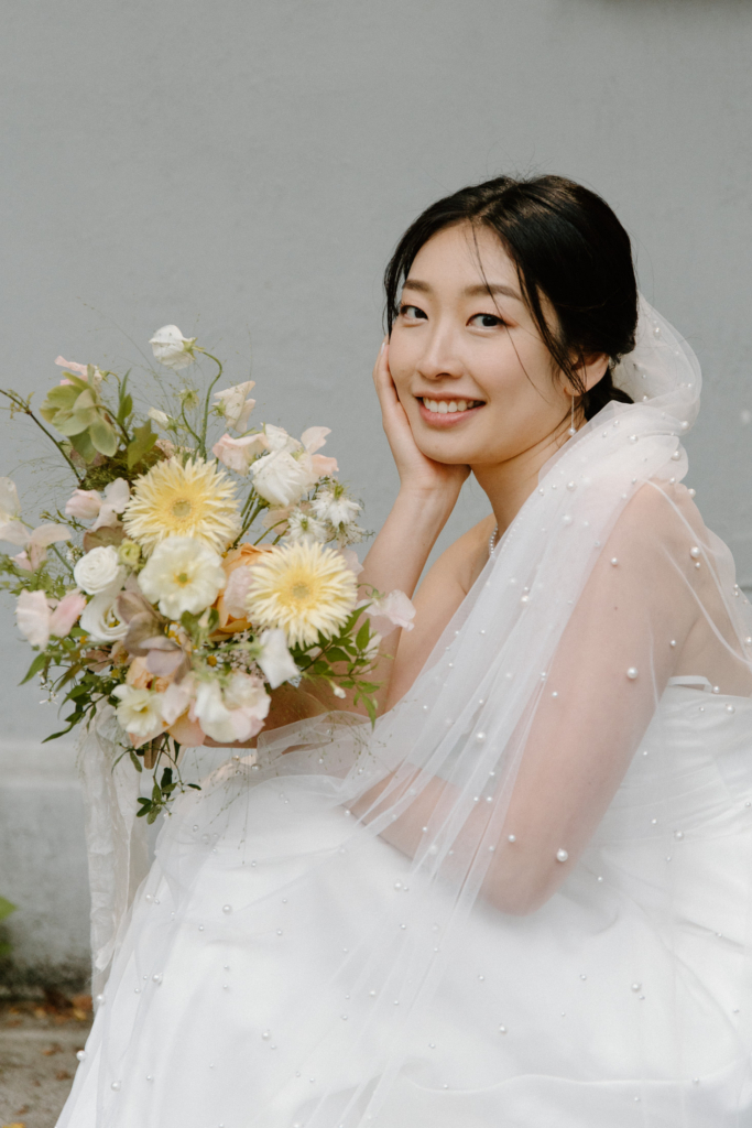 A bride sits in a soft, natural setting, smiling gently while holding a bouquet of light, pastel flowers. Her white dress and delicate veil with pearl details add texture, while the neutral background keeps the focus on her expression and the floral arrangement.