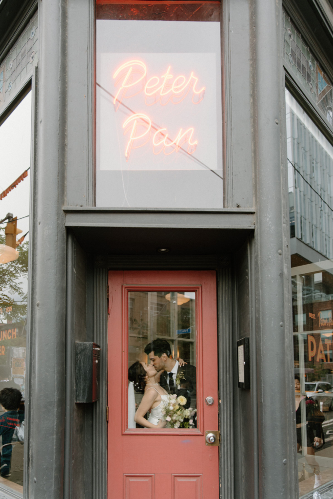 A bride and groom kiss inside the doorway of a city building, framed by a red door with a glass window. Above them, a glowing neon sign reads “Peter Pan,” adding a playful, urban contrast to the intimate wedding moment. Reflections of the street and passersby surround the scene, blending everyday life with the couple’s private moment.