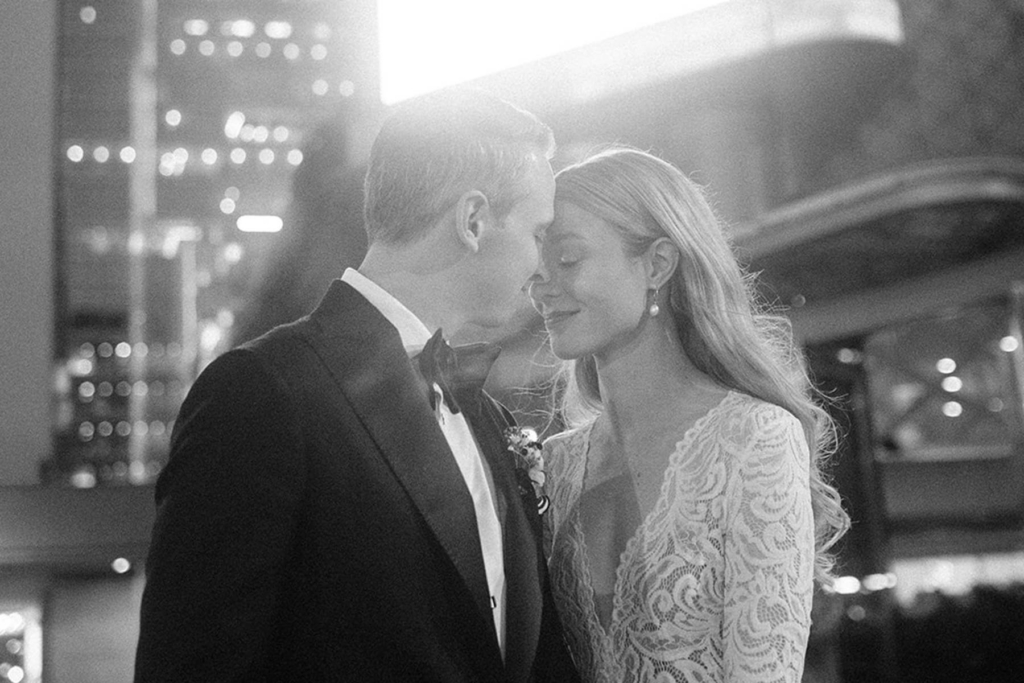 A bride and groom stand close together outdoors at night, their foreheads gently touching in a quiet, intimate moment. Soft city lights blur in the background, and the black-and-white tones emphasize the emotion and stillness between them.