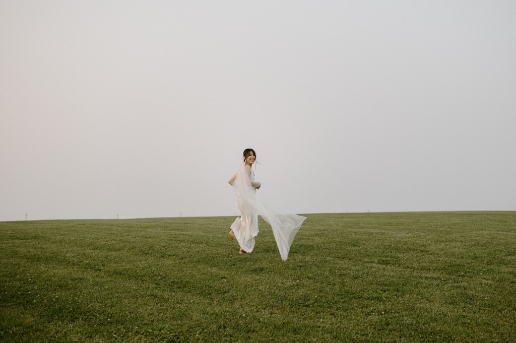 A bride runs across an open grassy field, her dress and veil moving in the wind under a wide, empty sky.