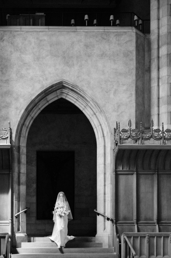 A bride in a veil walks down the steps of a large stone church, framed by a tall arched doorway in a black and white scene.