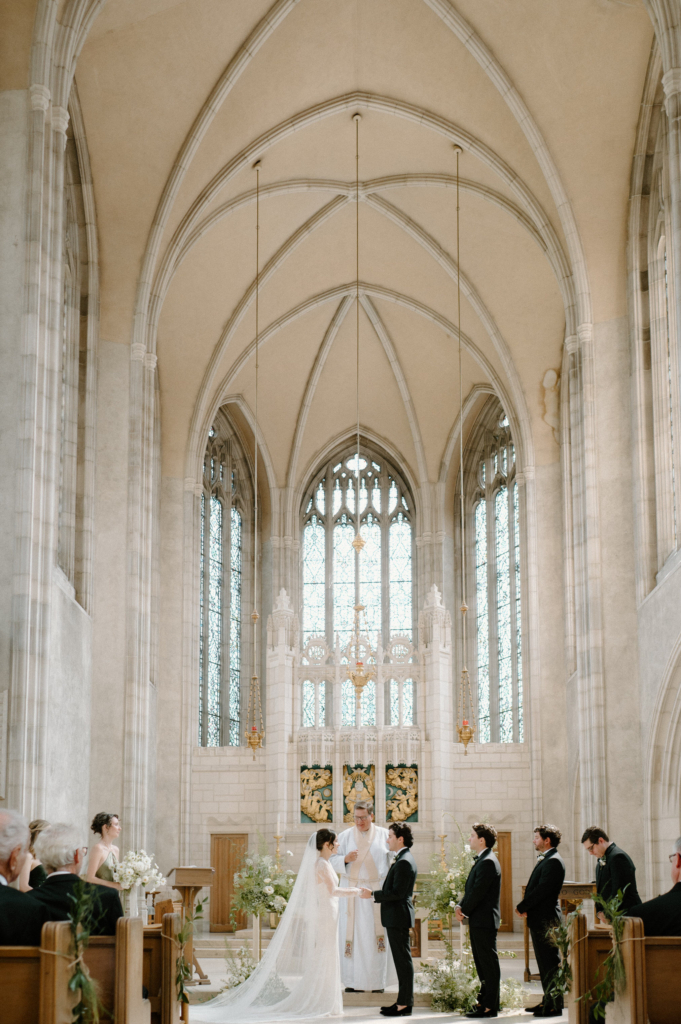 A couple stands at the altar during a wedding ceremony inside a bright church, surrounded by guests and tall arched windows.
