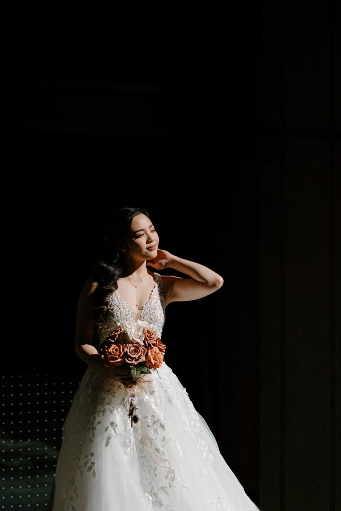 A bride stands in a dramatic beam of light against a dark background, holding a bouquet of warm-toned flowers. The contrast between light and shadow highlights the details of her dress and her calm, reflective expression. The scene feels cinematic and quiet, focusing attention on her presence within the surrounding darkness.