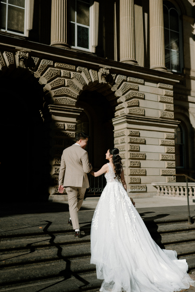 A bride and groom walk up stone steps toward a grand building, holding hands as sunlight casts strong shadows across the scene.
