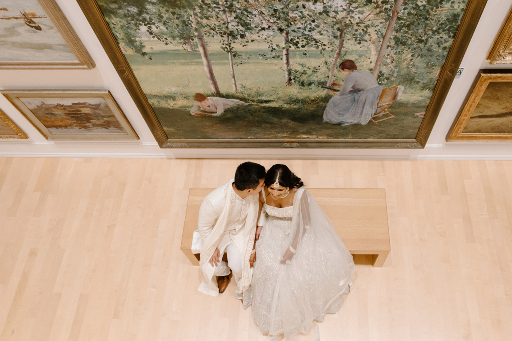 A couple sits closely together on a bench in an art gallery, surrounded by large paintings on the walls.