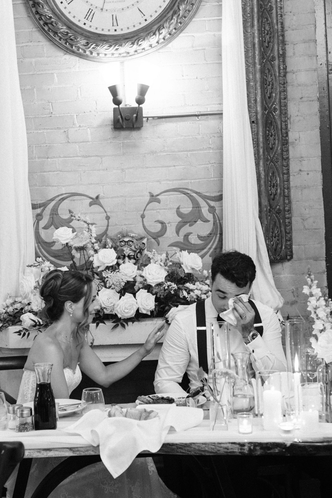 A groom sits at a wedding reception table, wiping tears from his face, while the bride gently touches his shoulder beside him.