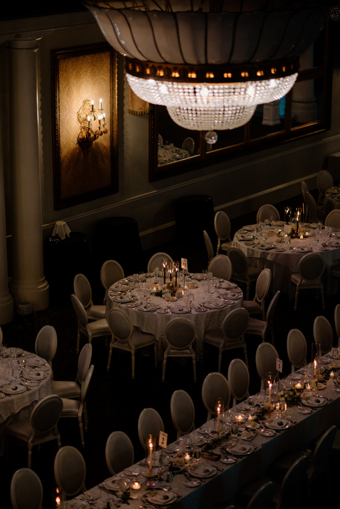 A warmly lit reception space is set with round tables and neatly arranged place settings, ready for guests. Soft candlelight and an elegant chandelier create a refined, intimate atmosphere. The scene captures the quiet anticipation before a wedding celebration begins.