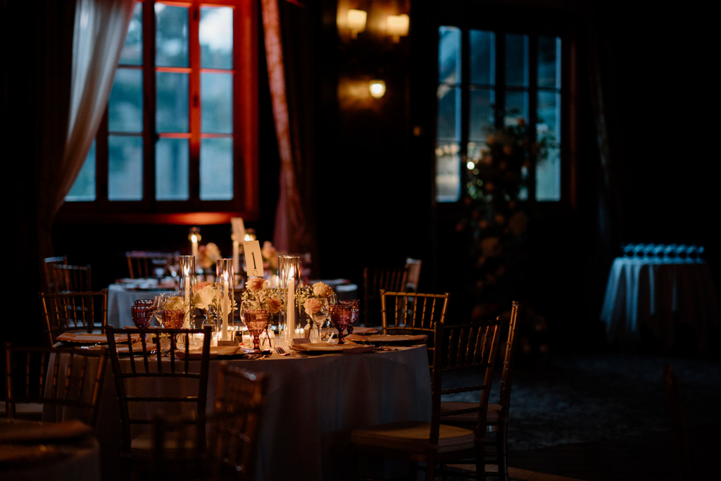A warmly lit wedding reception table set with candles, flowers, and glassware, arranged in an intimate indoor setting at night.