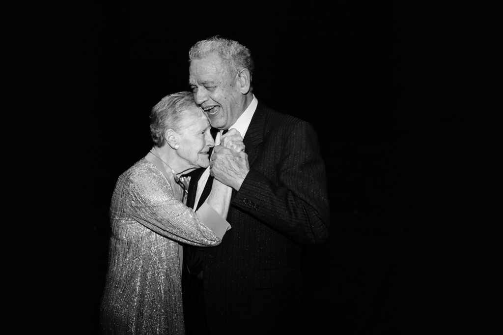 An elderly couple dances closely together against a dark background, smiling and holding each other with warmth and familiarity. The black-and-white image highlights their expressions and the tenderness of the moment, focusing on connection and shared history.