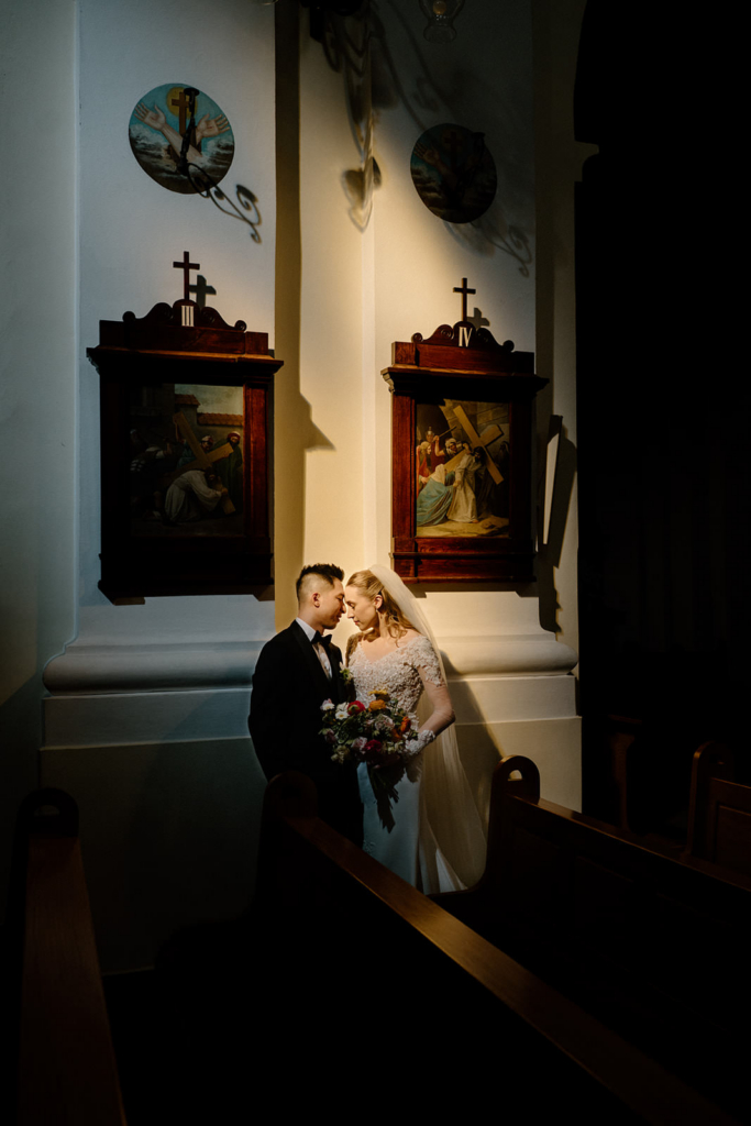 A bride and groom stand closely together inside a dimly lit church, their foreheads gently touching in an intimate moment. Soft, directional light falls on them, highlighting the bride’s white dress and bouquet while the surrounding space remains in shadow. Religious wall art and wooden pews frame the scene, creating a quiet, contemplative atmosphere around the couple.