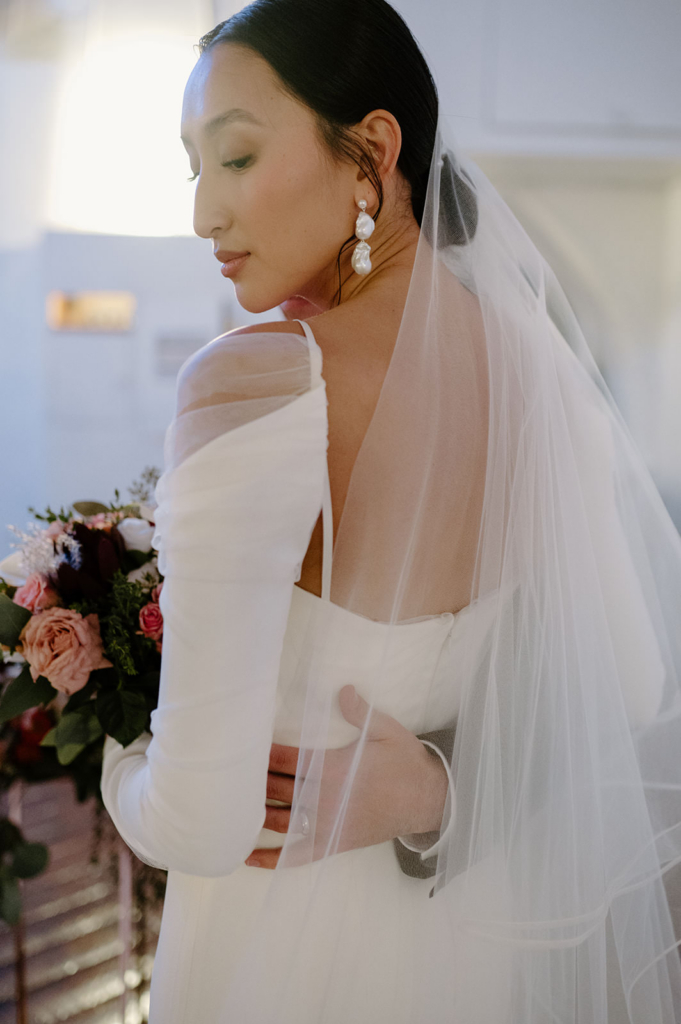 A bride is captured in profile, softly lit from behind, with a sheer veil flowing over her shoulders. She holds a bouquet of darker, richer-toned flowers, and her expression is calm and introspective, creating a quiet, elegant portrait.