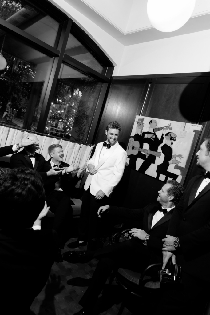 Black-and-white photograph of a groom in a white dinner jacket laughing with friends in tuxedos during a lively indoor wedding gathering.
Candid gestures, dramatic flash lighting, and dark wood interiors create an energetic, cinematic atmosphere.