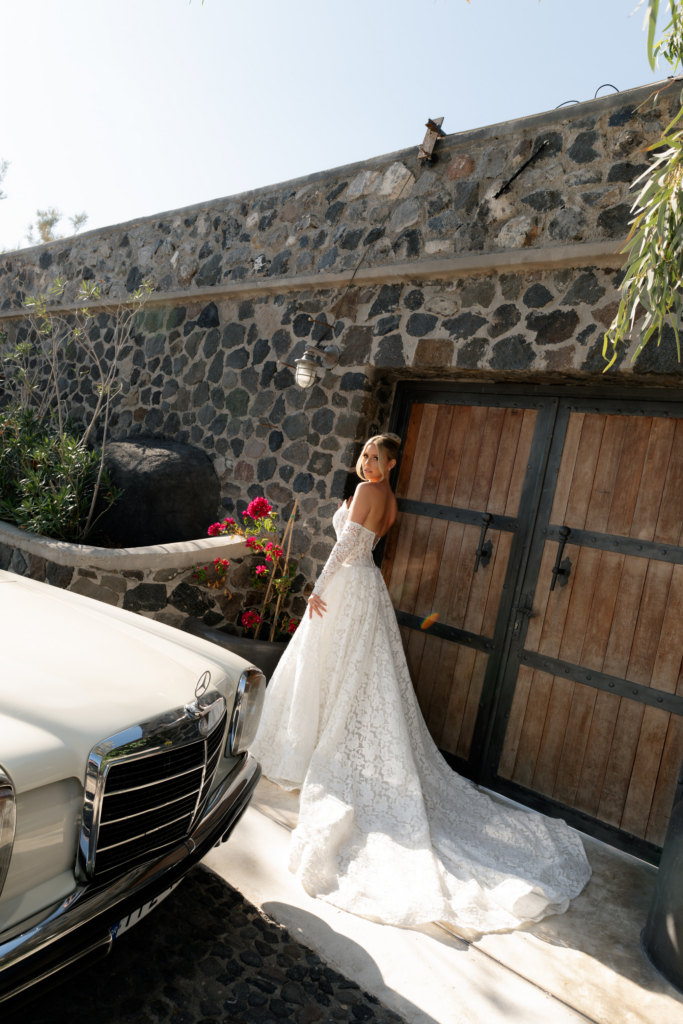 A bride in a long lace wedding gown stands beside a vintage white Mercedes in a sunlit stone courtyard.
Warm Mediterranean light, rustic textures, and flowing fabric create an elegant, cinematic portrait.