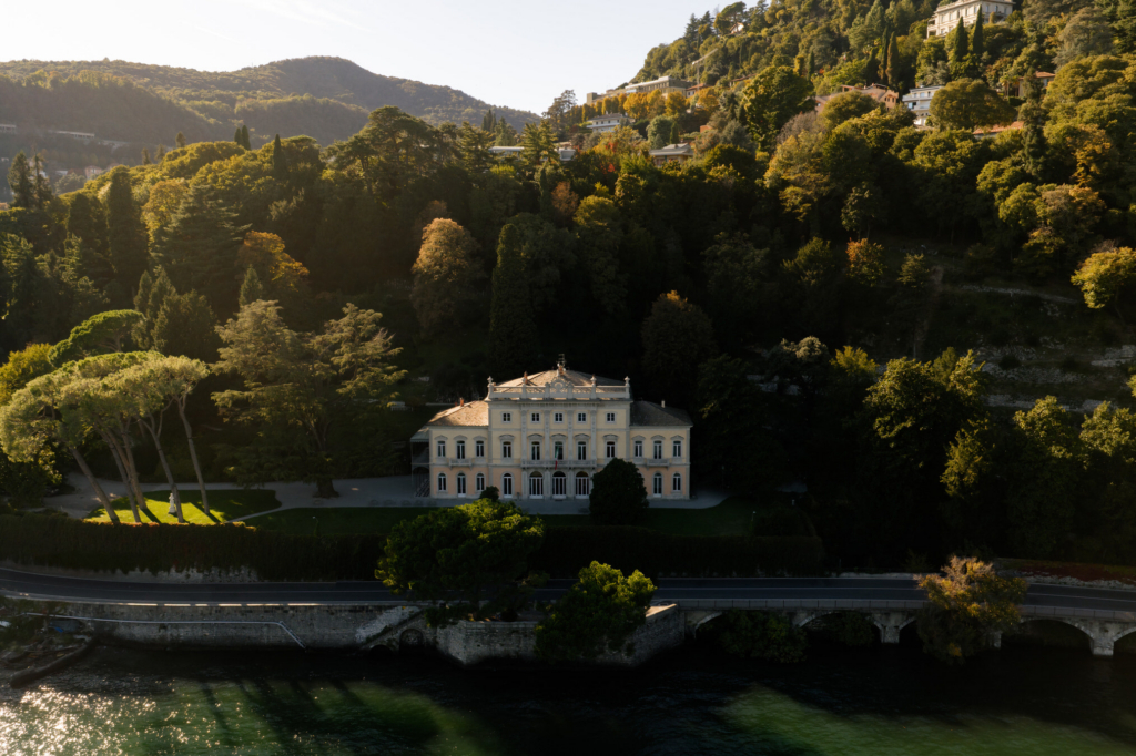 Aerial view of a grand lakeside villa surrounded by dense green hills and trees in warm evening light.
The symmetrical estate sits between the forested mountainside and the calm waterfront, creating a serene cinematic landscape.