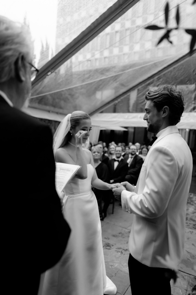 Black-and-white photograph of a bride and groom holding hands during their wedding ceremony as the bride wipes away tears.
Guests watch quietly in the background, capturing an intimate and emotional moment.
