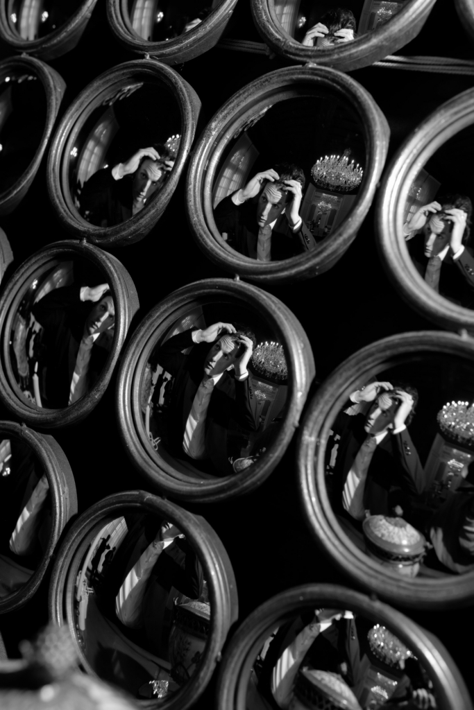 Black-and-white composition of a man adjusting his hair, reflected repeatedly in a wall of circular mirrors inside an elegant interior.
The layered reflections and dramatic lighting create a surreal, cinematic portrait with a sense of repetition and movement.