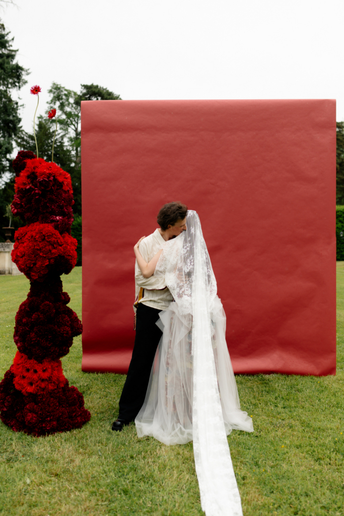 A couple embracing beneath a long lace veil in front of a deep red backdrop set outdoors on a grassy field.
The composition combines theatrical color, soft textures, and an intimate moment between the two figures.