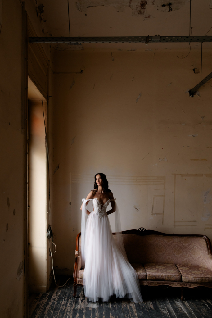 A bride in a flowing tulle gown stands beside a vintage sofa inside a worn, dimly lit room with peeling walls and wooden floors.
Soft natural light and the large empty space create a quiet, cinematic mood with an editorial feel.