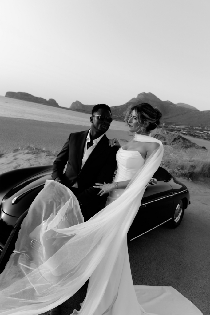 Black-and-white portrait of a couple posed beside a vintage convertible on a coastal road, with mountains and sea in the background.
Flowing fabric and soft evening light create a cinematic sense of movement and intimacy.