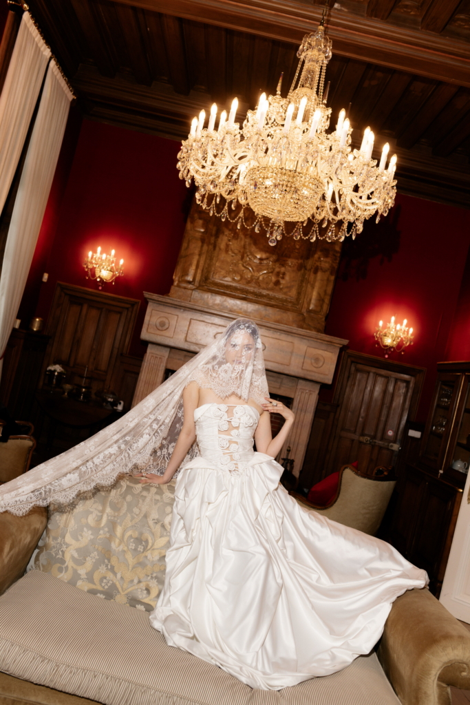 A bride in a sculptural satin gown and long lace veil poses beneath crystal chandeliers in an ornate historic interior.
Rich red walls, dark wood details, and dramatic lighting create a regal, theatrical atmosphere.
